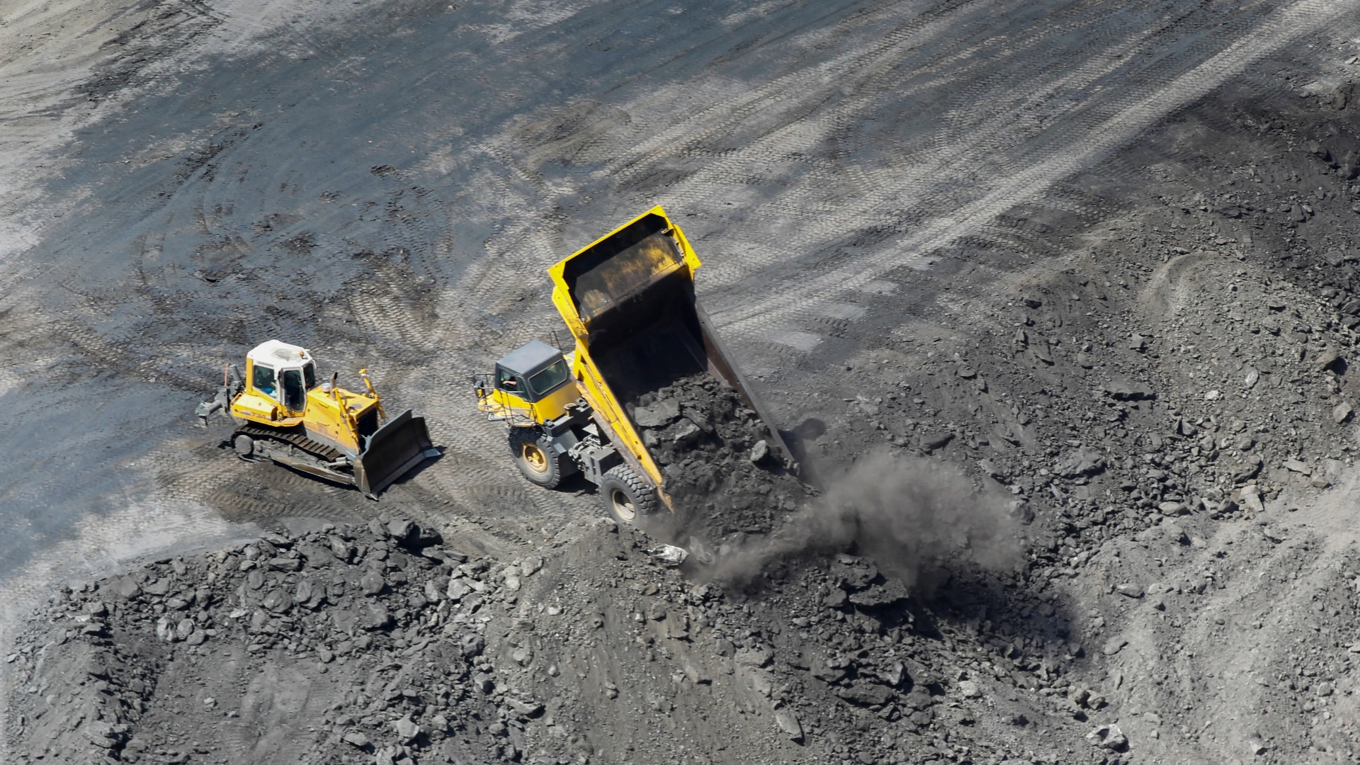 Großhandel mit Kohle — Draufsicht auf einen gelben Kipper, der Kohle in eine große Grube entlädt, daneben gleicht ein Bulldozer das Gelände auf einem industriellen Bergbaugelände aus.